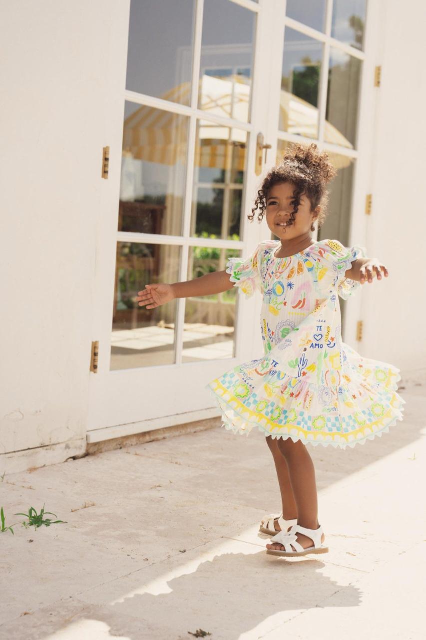 Girl twirling in a white printed dress with a scalloped hem outside a sunlit doorway